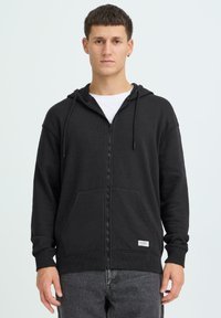 Sweat à capuche zippé noir en tissu doux, avec capuche, deux poches avant et poignets côtelés. Étiquette avec logo en accent sur l'ourlet.