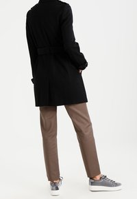 Zwarte trenchcoat met een tailleband, gecombineerd met bruine broeken en grijze sneakers. De jas heeft een klassiek ontwerp met een gestructureerde silhouette.