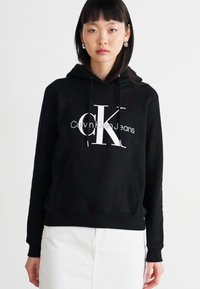 Juodas gobtuvas iš minkštos medvilnės. Puikuojasi dideliu baltu Calvin Klein logotipu su "CK" moderniu šriftu. Paprastas dizainas ir atsipalaidavęs siluetas.