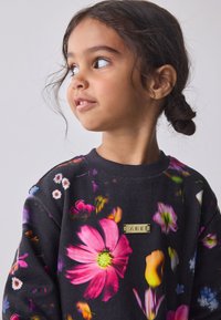 Mustast riidest pusakast, millel on lillemuster roosades, lillades ja valgetes toonides ning kuldse logoga esiküljel.