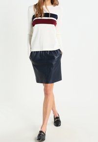 Långärmad krämfärgad topp med marinblå och burgundy-ränder, kombinerad med en marinblå läderkjol. Svarta loafers med guldfärgad metallaccents.