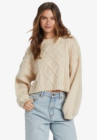 Pull en tricot crémeux avec des motifs en losanges, manches larges et ourlet côtelé, associé à un jean bleu clair taille haute.
