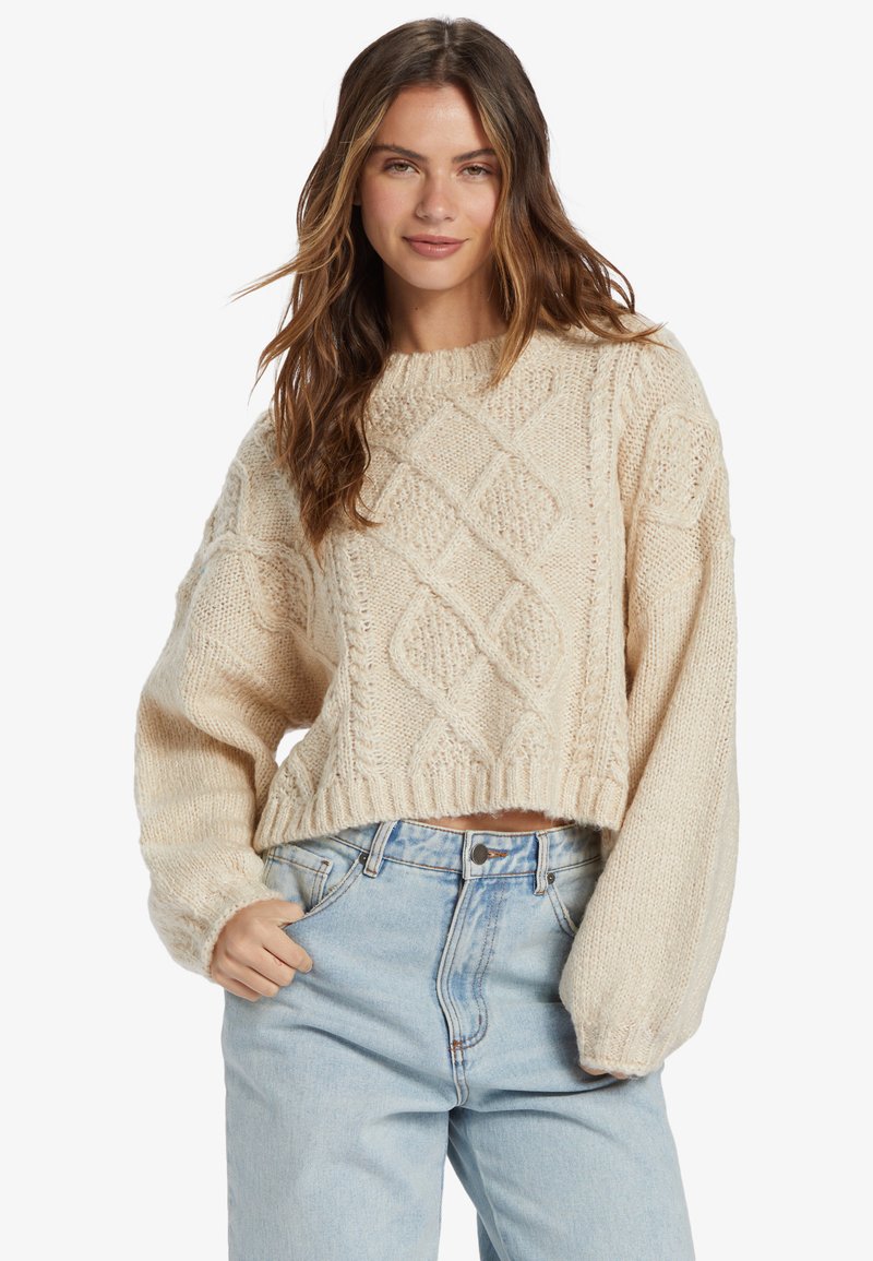 Pull en tricot crémeux avec des motifs en losanges, manches larges et ourlet côtelé, associé à un jean bleu clair taille haute.
