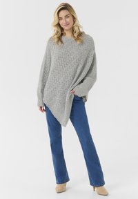 Ljust grå oversized tröja med ett zickzackstickat mönster, vida ärmar och en asymmetrisk nederkant, kombinerad med blå raka jeans och beige klackar.