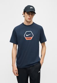 Νεαρός άνδρας που φοράει μπλε ναυτικό μπλουζάκι Nike με εξαγωνικό λογότυπο και μαύρο καπέλο και παντελόνι Nike που ταιριάζουν, στέκεται με το ένα χέρι στην τσέπη.