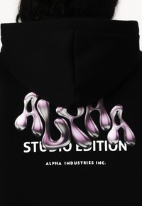 Čierna mikina s kapucňou s trojrozmerným grafickým dizajnom "ALPHA" v fialovej a striebornej farbe a textom "STUDIO EDITION" v bielej farbe nižšie. Hladká tkanina s kapucňou.