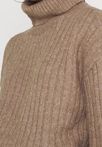 Beige värillinen ribbineulottu neulepaita, jossa on korkea poolokaulus, pehmeästä materiaalista valmistettu, joka näyttää hienovaraisen rakenteen ja istuvan siluetin.