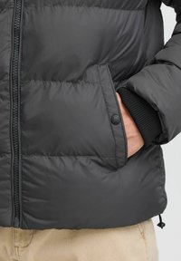 Sort pufferjakke i materiale med quiltet design, der har en lynlås foran, sidelomme og ribbede manchetter. Glat tekstur og minimal detaljering.