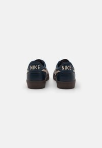 Tamsiai mėlynos Nike sportinės bateliai su lygiąja odos danga, apvalia nosimi ir smėlio spalvos akcentais. Yra iškilus logotipas ant kulno ir guminis padas.