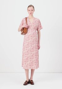 Rochie maxi florală în roz deschis, cu mâneci scurte, talie strânsă și tiv evazat. Asortată cu o geantă de umăr maronie și loafers negri.