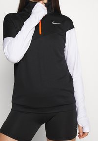 Mustavalkoinen pitkähihainen urheilu paita korkealla kauluksella, oranssi vetoketju yksityiskohtana ja Nike-logo, yhdistettynä mustiin shortseihin.