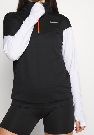 Mustavalkoinen pitkähihainen urheilu paita korkealla kauluksella, oranssi vetoketju yksityiskohtana ja Nike-logo, yhdistettynä mustiin shortseihin.