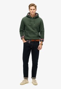 Felpa verde con cappuccio e tasca a marsupio, indossata sopra una camicia di flanella a check; abbinata a jeans scuri e sneaker beige.