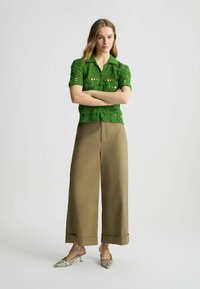 Camicetta verde all'uncinetto a maniche corte con colletto, abbinata a pantaloni beige a gamba larga. L'outfit presenta una vestibilità ampia e una texture a motivo.