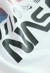 Tricou alb din material textil, cu un text mare negru și desene grafice, inclusiv forme geometrice și o ilustrație cu un personaj de desene animate.