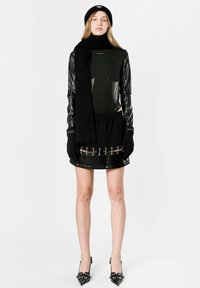 Top negro de manga larga con mangas de cuero brillantes, bufanda negra en capas, falda negra ajustada con herrajes plateados y detalles transparentes, tacones altos.