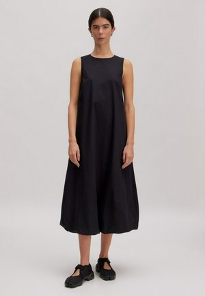 Vrouw staand in een mouwloze, losse zwarte midi-jurk en zwarte wikkel schoenen tegen een effen lichte achtergrond.