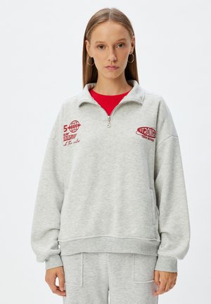 Sudadera de cremallera gris claro con cuello alto, que presenta texto y gráficos impresos en rojo. Fabricada con un tejido suave y texturizado. Mangas largas.
