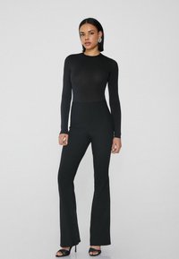 Černý body s dlouhým rukávem v kombinaci s vysokými rozšířenými kalhotami, obojí z hladkého, elastického materiálu. Modelka má na sobě černé sandály na podpatku.