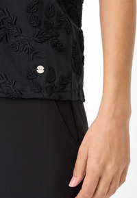 Sort top med intrikate tonale broderier af blade, med en lille sølvlogo-detalje og en glat stoftekstur.