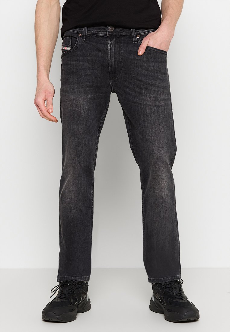 Mann in abgetragenen schwarzen, gerade geschnittenen Jeans und schwarzen Sneakers mit einer Hand in der Tasche vor einem einfarbigen Hintergrund.