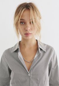 Camicia a righe bianca e nera, con colletto. Il modello indossa una collana in argento. Capelli acconciati in modo sciolto. Sfondo chiaro.