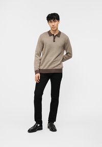 Νέος άνδρας που στέκεται με το ένα χέρι πίσω από την πλάτη, φορώντας μπεζ πουλόβερ πόλο με μακριά μανίκια, μαύρο παντελόνι και μαύρα παπούτσια, σε απλό λευκό φόντο.