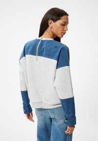 Felpa grigia chiara con dettagli blu. Maniche lunghe con bottoni, colletto rotondo e chiusura con zip sul retro. Materiale morbido e vestibilità comoda.
