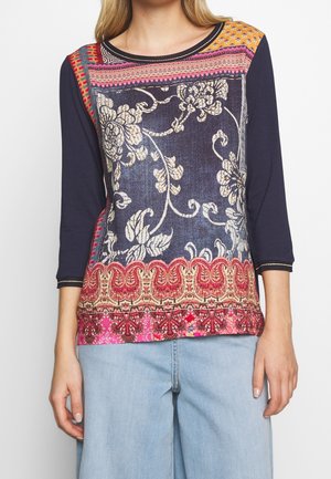 Femme portant un haut à manches longues bleu marine avec des motifs floraux et géométriques multicolores, associé à un jean bleu clair.