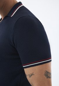Marineblaues Polohemd mit kurzen Ärmeln und strukturiertem Muster, mit einem Kragen, dessen Ränder rot und weiß gestreift sind.