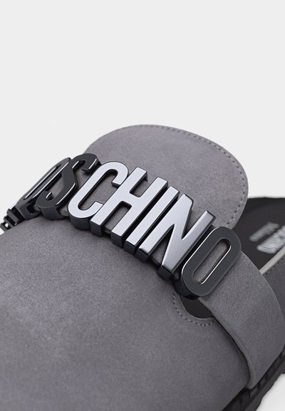 Γκρι παπούτσι χωρίς κορδόνια με μεγάλα μαύρα και λευκά ανάγλυφα γράμματα που σχηματίζουν τη λέξη "MOSCHINO" στο λουρί στην πάνω πλευρά του ποδιού.