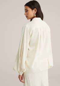 Vrouw met lang donker haar, die gouden oorbellen draagt en een ivoorkleurige blouse met lange geplooide mouwen en een plooi aan de achterkant.