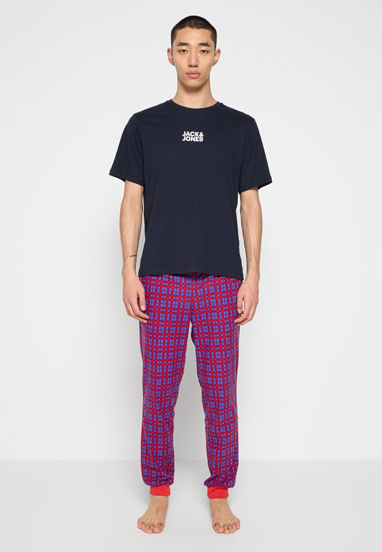 Joven de pie descalzo, vistiendo una camiseta negra de Jack & Jones y pantalones de pijama a cuadros rojos y azules.