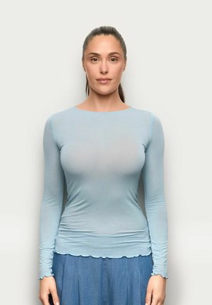 Kobieta z włosami związanymi do tyłu, ubrana w dopasowany jasnoniebieski top z długim rękawem i niebieską spódnicę jeansową, stoi na tle jednolitego białego tła.