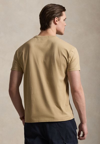 Μπεζ βαμβακερό μπλουζάκι με κοντά μανίκια, με στρογγυλή λαιμόκοψη, που φοριέται από άτομο που φαίνεται από πίσω, συνδυασμένο με σκούρο σορτς.