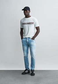 Άνδρας που φοράει λευκό T-shirt Dsquared2, ανοιχτό μπλε τζιν, μαύρο καπέλο και μαύρα αθλητικά παπούτσια, στέκεται με τα χέρια στις τσέπες μπροστά σε απλό φόντο.