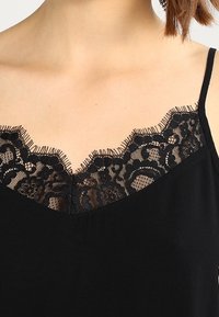 Kvinna som bär en svart linne med intrikata blommönster i spets längs halsringningen och tunna axelband.