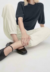 Personne assise en tailleur portant un haut noir à manches courtes en maille, un pantalon crème à poignets élastiques et des chaussures Mary Jane noires.