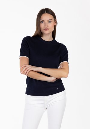 Vrouw met lang bruin haar, gekleed in een marineblauw shirt met korte mouwen en een witte broek, staat met gekruiste armen tegen een witte achtergrond.