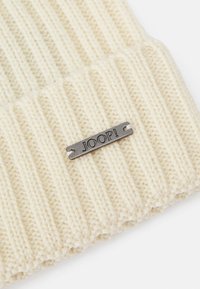 Berretto a maglia crema con texture a coste, dotato di un'etichetta grigio metallico con la scritta "JOOP!" in rilievo; materiale morbido e caldo, ideale per il freddo.