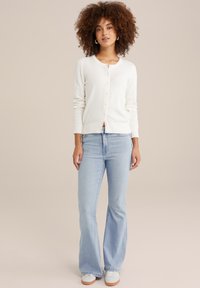 Bijela teksturirana cardigan s prednjim gumbima, uparen s svijetloplavim flare trapericama i bijelim tenisicama. Model stoji naslonjen na neutralnu pozadinu.