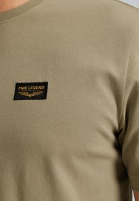 Camisa de algodón color verde oliva con mangas cortas, que presenta una etiqueta negra rectangular con texto dorado y un diseño de alas en el pecho.