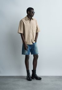 Hombre con camisa de manga corta a cuadros, pantalones cortos de mezclilla, botas negras hasta la mitad de la pantorrilla y gafas de sol, de pie contra una pared gris lisa.