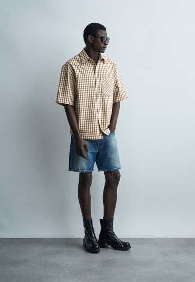 Homme portant une chemise à manches courtes à carreaux, un short en denim, des bottes noires mi-mollet et des lunettes de soleil, debout contre un mur gris uni.