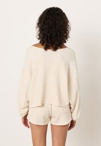 Šviesiai smėlio spalvos laisvo stiliaus megztinis su nusmukusiomis pečių linijomis ir plačia iškirpte. Derinamas su minkštais, tekstūruoto audinio šortais, abi dalys pagamintos iš tos pačios medžiagos.