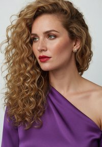 Top viola con spalle scoperte e texture liscia, abbinato a orecchini d'oro a cerchio. La modella ha capelli ricci biondi e labbra rosse intense.