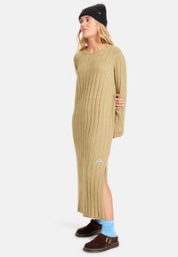 Maxikleid aus Khaki mit vertikalen Rippen, seitlichem Schlitz und Logo-Akzent. Kombiniert mit blauen Socken und braunen Clogs, garniert mit einer schwarzen Mütze.