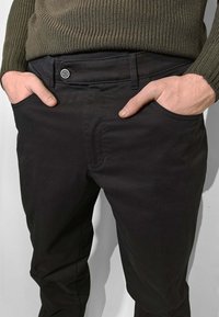 Pantaloni chino neri in cotone con fronte piatto, due tasche laterali, chiusura con bottone e design slim-fit. Vita testurizzata in verde oliva.