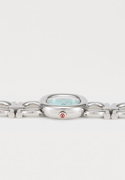 Reloj de metal plateado con esfera redondeada, esfera verde menta y botón rojo destacado. Presenta un diseño de correa con cadena enlazada.