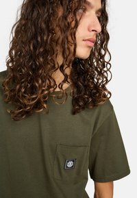 Olive green t-shirt made of cotton, featuring a chest pocket with a black label. Short sleeves and a relaxed fit.